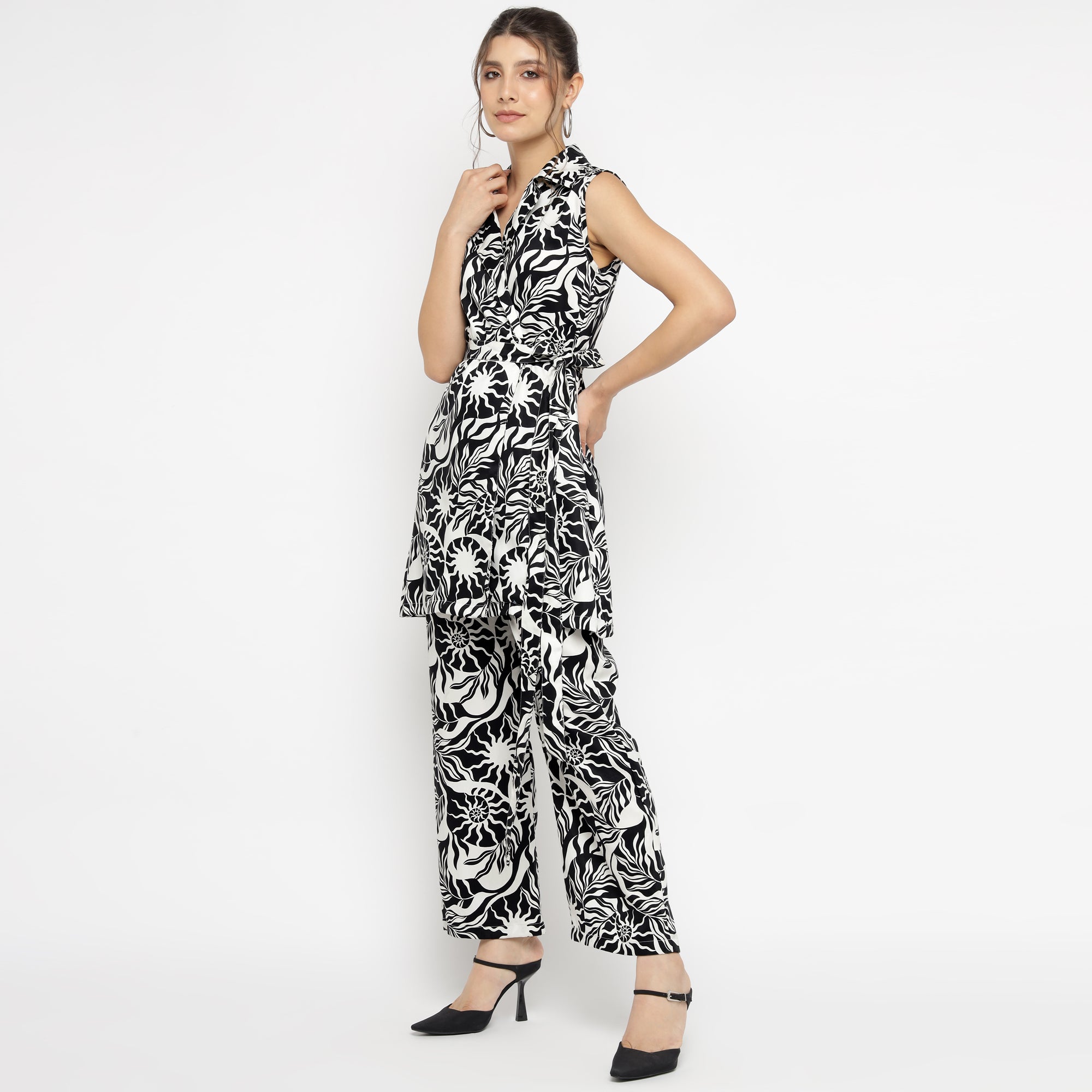 Model posing in a sleeveless black and white abstract printed co-ord set, featuring a collared V-neckline top with an asymmetrical hem and matching wide-leg pants. Styled with black heels and hoop earrings, set against a plain white backdrop.